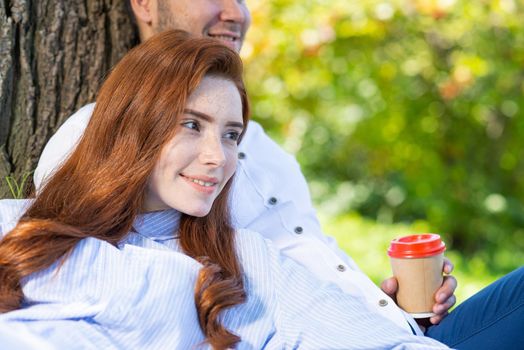 Young couple relaxing under tree in summer park on sunny day. Happy couple in love spend time outdoors together. Handsome man and pretty redhead girl enjoying each other. Romantic relationships.