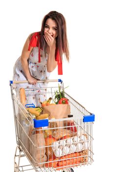 A young female pushing a shopping cart full with groceries isolated on white background. Brunette in a summer dress makes purchases. A woman laughs and closes her mouth with her palm