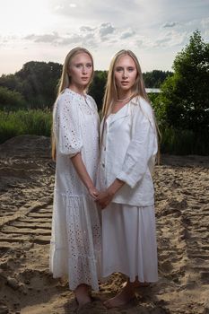young pretty twins with long blond hair posing at sand beach in elegant white dress, skirt, jacket. stylish summer fashion photoshoot with flashlight. identical sisters spend time together outdoors