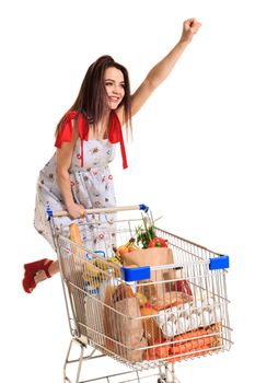 Woman with shopping cart full with products isolated over white background. A young woman rides for fun on an iron shopping basket, raising her hand up like a superman