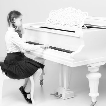 Charming little blonde with long wattled hair in plaits, playing on a white grand piano. In musical school.Black-and-white photo. Retro style.