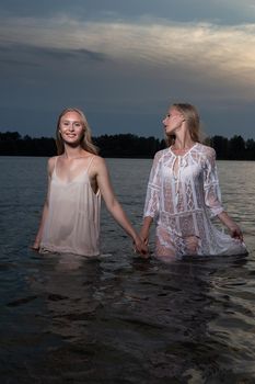 two attractive young twin sisters with long blond hair posing in light dresses in water of lake at summer night. stylish fashion photoshoot with flashlight. pretty models outdoors evening photosession