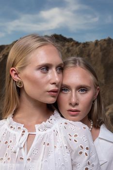 two young blond pretty twins posing at sand quarry in elegant white clothes. stylish glamorous fashion photoshoot with flashlight. girl holds her head on shoulder of her sister in front of sand wall