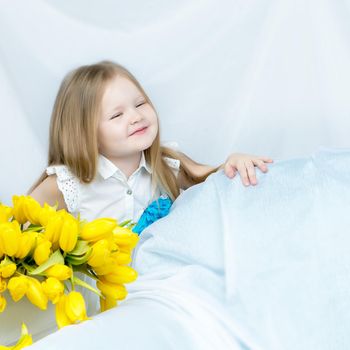 Cute little girl with long blond hair in a long blue skirt sitting on the couch with a large bouquet of bright yellow tulips.