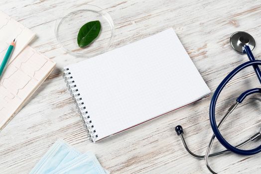 Patient examination and consultation concept with doctor workplace. Spiral notepad, disposable face mask, stethoscope and cardiogram on wooden surface. Professional healthcare in medical center.