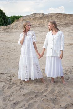 two young pretty twins with long blond hair posing at sand quarry in elegant white dress, skirt, jacket. stylish fashion photoshoot, summer photosession. identical sisters spend time together outdoors