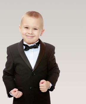 Beautiful little blond boy in a fashionable black suit with a tie.The boy tightly clenched his fists.On a gray background.