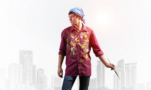Young artist posing with paintbrush. Painter in dirty shirt and bandana standing on background modern downtown. Creative hobby and artistic occupation concept with copy space. Art classes advertising
