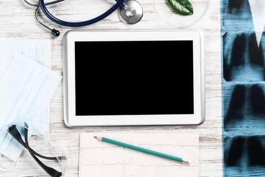 Flat lay workplace of doctor in modern clinic. Tablet computer with blank screen, x-ray image, stethoscope and cardiogram lies on wooden desk. Professional medical consultation and treatment