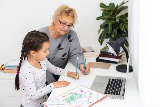 Mature grandmother helping child with homework at home. Satisfied old grandma helping her granddaughter studying in living room. Little girl writing on notebook with senior teacher sitting next to her