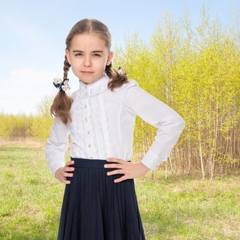 A beautiful little schoolgirl girl in a white blouse and black long skirt, with neatly braided pigtails on her head.She is posing in front of the camera, her hands on her hips.Against the background of the summer landscape.