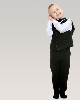 Beautiful little blond boy in a fashionable black suit with a tie.He keeps his hands near your ear.On a gray background.