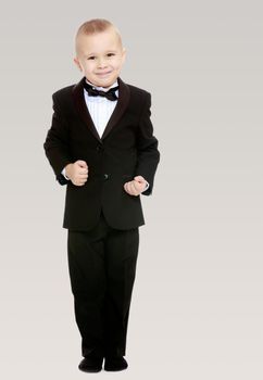 Beautiful little blond boy in a fashionable black suit with a tie.The boy tightly clenched his fists.On a gray background.