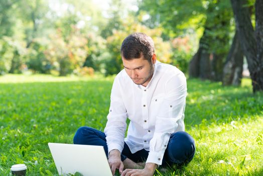 Young businessman sitting on green grass and using laptop computer. Handsome man working with computer in park at sunny summer day. Outdoors nature journey and relaxation. Freelance work concept.