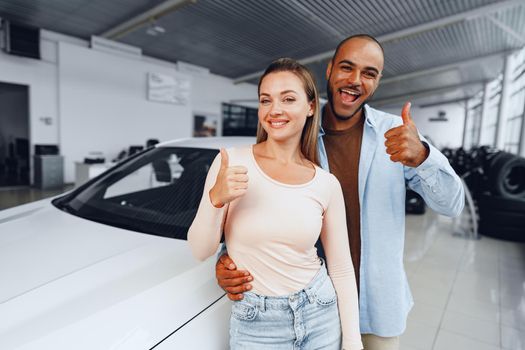 Happy couple of caucasian woman and african american man standing near their new luxury car inside car salon