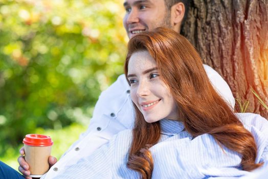 Young couple relaxing with coffee under tree in park on sunny day. Happy couple in love spend time outdoors together. Handsome man and pretty redhead girl sitting on green grass leaning against tree