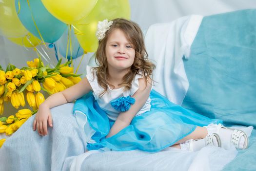 Beautiful little girl in a blue skirt tutu, sitting on the couch with a bouquet of yellow tulips.