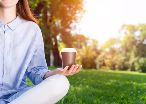Young redhead woman meditating in lotus pose on green grass. Close up woman hand holding take away coffee cup. Girl in casual wear practicing of yoga in park. Mental relax and balance concept.