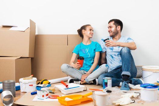 Happy smiling couple relaxing on floor with cup of coffee among construction tools and materials. House remodeling and interior renovation concept. Young man and woman together painting wall at home.