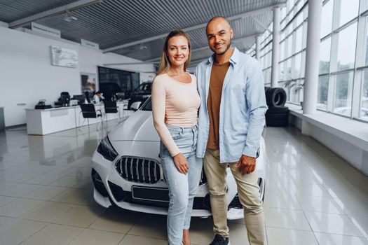 Happy couple of caucasian woman and african american man standing near their new luxury car inside car salon