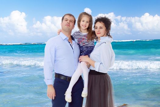 Happy young family, mom dad and little daughter.Parents keep the girl in her arms , and she hugs their neck.White sand , blue sea and blue sky with clouds.