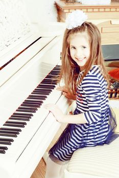 Little girl in blue striped dress and a white bow on her head.Girl smiling sitting behind the keys of a large white piano.Creative toning of a photograph.