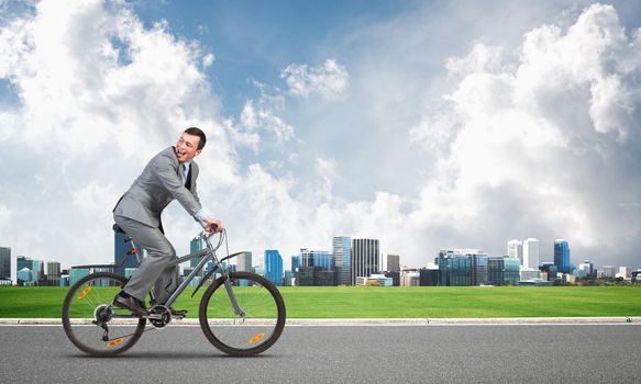 Businessman commuting to work by bike. Man wearing business suit riding bicycle on asphalt road. Handsome cyclist looking back on background of modern cityscape and blue cloudy sky.