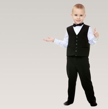 Beautiful little blond boy in a fashionable black suit with a tie.He shows his hand to the side.On a gray background.