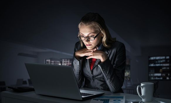 Portrait of concentrated businesswoman in glasses sitting at office desk and working at laptop. Financial consultant in business suit and tie busy at work in evening. Working in office late at night.