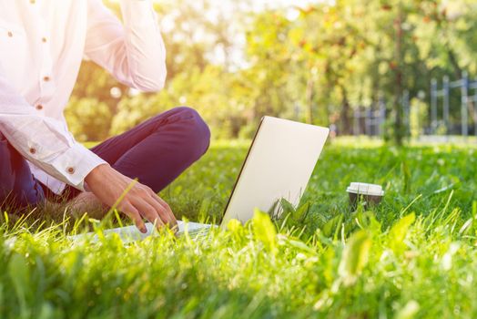 Businessman using laptop computer on green grass in park. Close up man hands typing on keyboard. Man in casual wear working with computer outdoors at sunny summer day. Freelance and blogging concept.