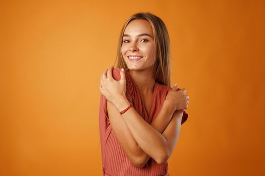 Peaceful young blonde woman holding hands on her chest against orange background.