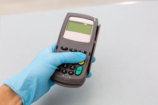 Cashier hand holding credit card reader machine and wearing disposable gloves, paying with smartphone during Covid-19 pandemic.