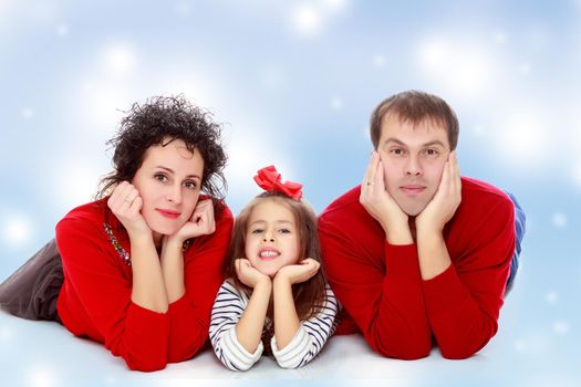 Happy young family dad mom and a little girl in bright red outfits . Family lying on the floor leaning on his hands.Blue Christmas festive background with white snowflakes.