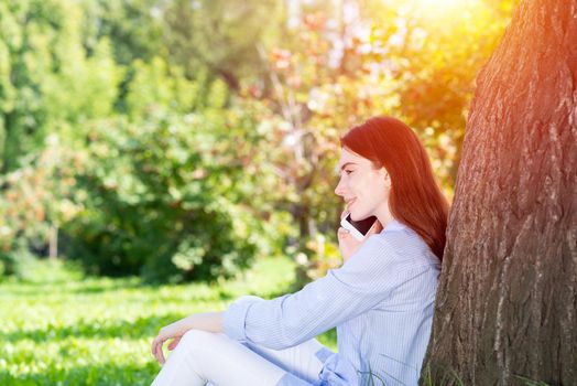 Young redhead woman talking on smartphone under tree in park on sunny day. Beautiful girl relaxing outdoors. Charming girl sitting on green grass leaning against tree. Mobile communication concept.