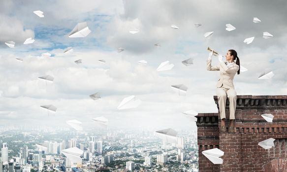 Young woman playing trumpet and sitting on roof. Girl in white business suit with music brass instrument posing high above modern city. Imagination and inspiration. Flying paper planes in blue sky