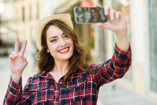 Portrait of a beautiful young woman selfie in the street with a smartphone. Young woman wearing plaid shirt and sun hat with red lips in urban background.