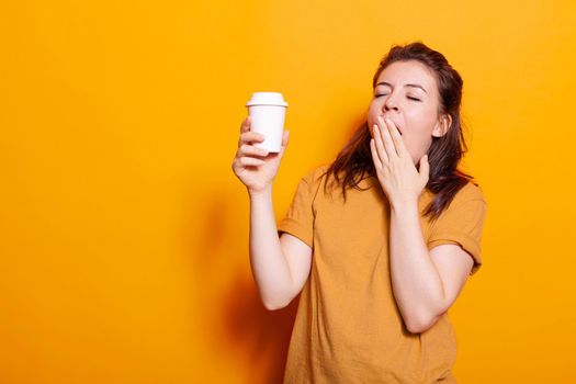 Sleepy woman yawning while holding cup of hot coffee on camera. Young person feeling exhausted and falling asleep while drinking beverage with caffeine to stay awake and have energy.