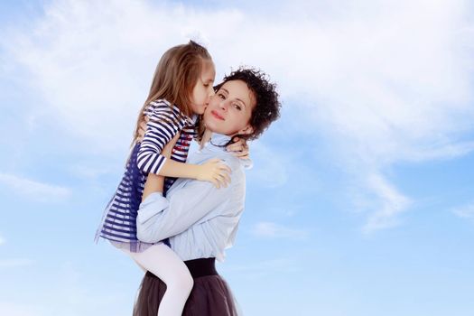 Happy young beautiful mother,lifted his little beloved daughter .And the girl kisses his mother on the cheek.On the background of summer blue sky and fluffy clouds.