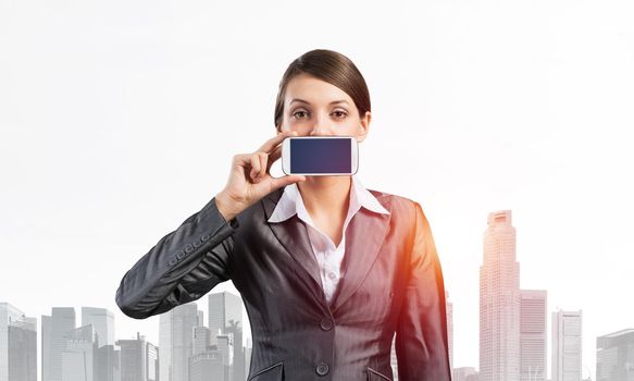 Portrait of young woman covering her mouth with smartphone. Businesswoman showing mobile phone with blank screen. Corporate businessperson on cityscape background. Mobile communication layout.