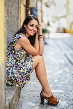 Happy young woman with blue eyes smiling sitting on urban step. Girl wearing flower dress in urban background. Beauty and fashion concept.
