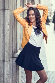 Young brunette woman, model of fashion, wearing orange modern jacket and blue skirt. Pretty caucasian girl with long wavy hairstyle smiling. Female raising her arms in urban background.