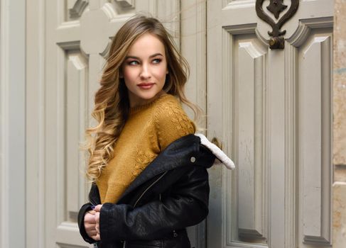 Blonde woman in urban background. Beautiful young girl wearing black leather jacket and mini skirt standing in the street. Pretty russian female with long wavy hair hairstyle and blue eyes.