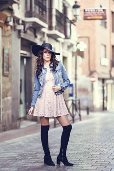Young brunette woman, model of fashion, wearing denim jacket, hat, long socks and pink dress. Pretty caucasian girl with long wavy hairstyle in urban background.