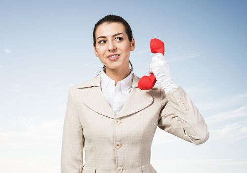 Attractive young woman holding red retro phone. Call center operator in white business suit posing with telephone on skyscape background. Hotline telemarketing. Business assistance and consultation.