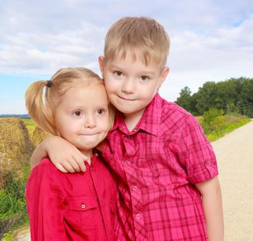 The concept of family happiness and children , against the background of fresh green grass, mown hay stacks, and a dirt road receding into the distance.Little brother and sister cute hug.