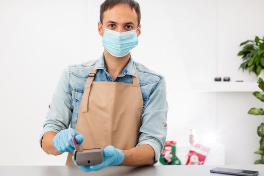 Cashier hand holding credit card reader machine and wearing disposable gloves, paying with smartphone during Covid-19 pandemic.