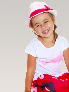 Widely smiling a small, tanned girl in the pink hat sits on the floor,folding his Turkish legs. On a gray background.