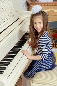Little girl in blue striped dress and a white bow on her head.Girl smiling sitting behind the keys of a large white piano.