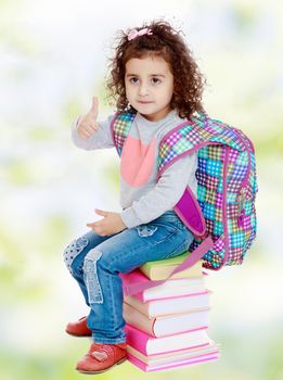 Beautiful little curly girl in jeans and t-shirt sitting on a stack of books. The girl gestures raising his thumb up.white-green blurred abstract background with snowflakes.