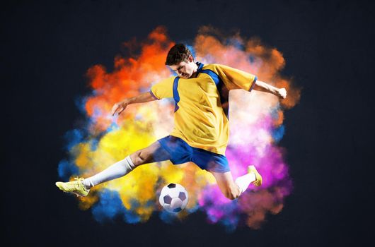 Soccer player kicking ball in colorful smoke. Sportsman in yellow and blue uniform in action. Soccer game championship concept with copy space. Young player on dark background. Movement at gameplay.